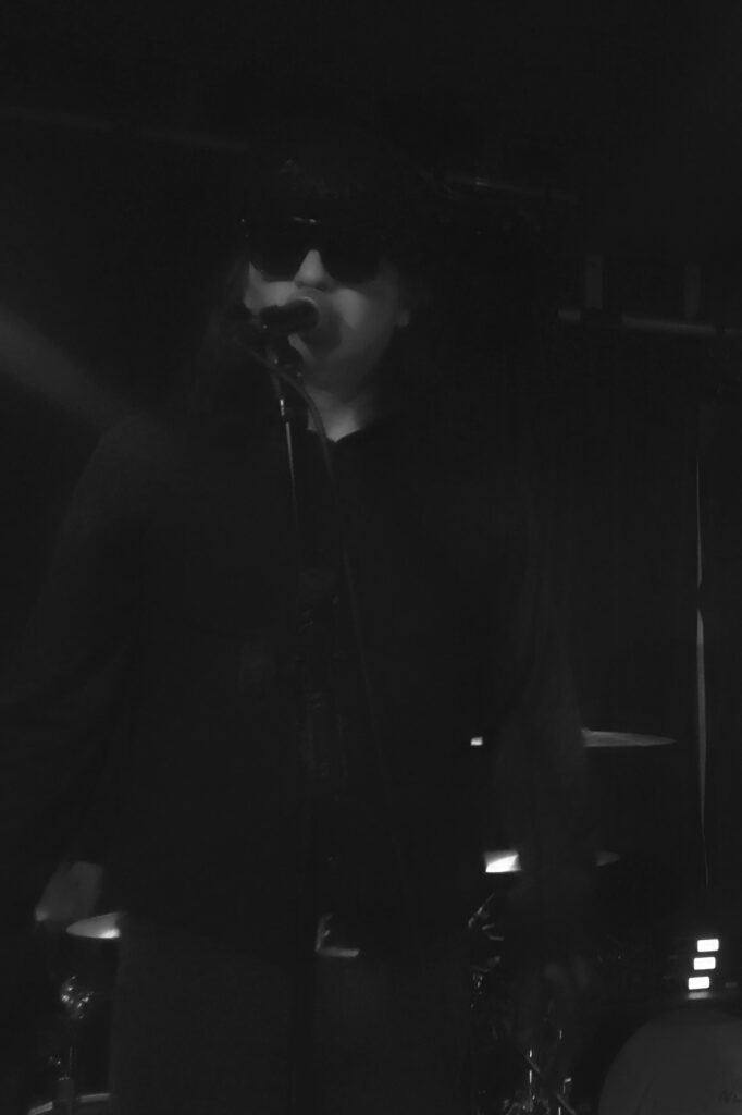 Mann mit Sonnenbrille und schwarzer Kleidung, der auf einer Bühne singt und ein Mikrofon hält.