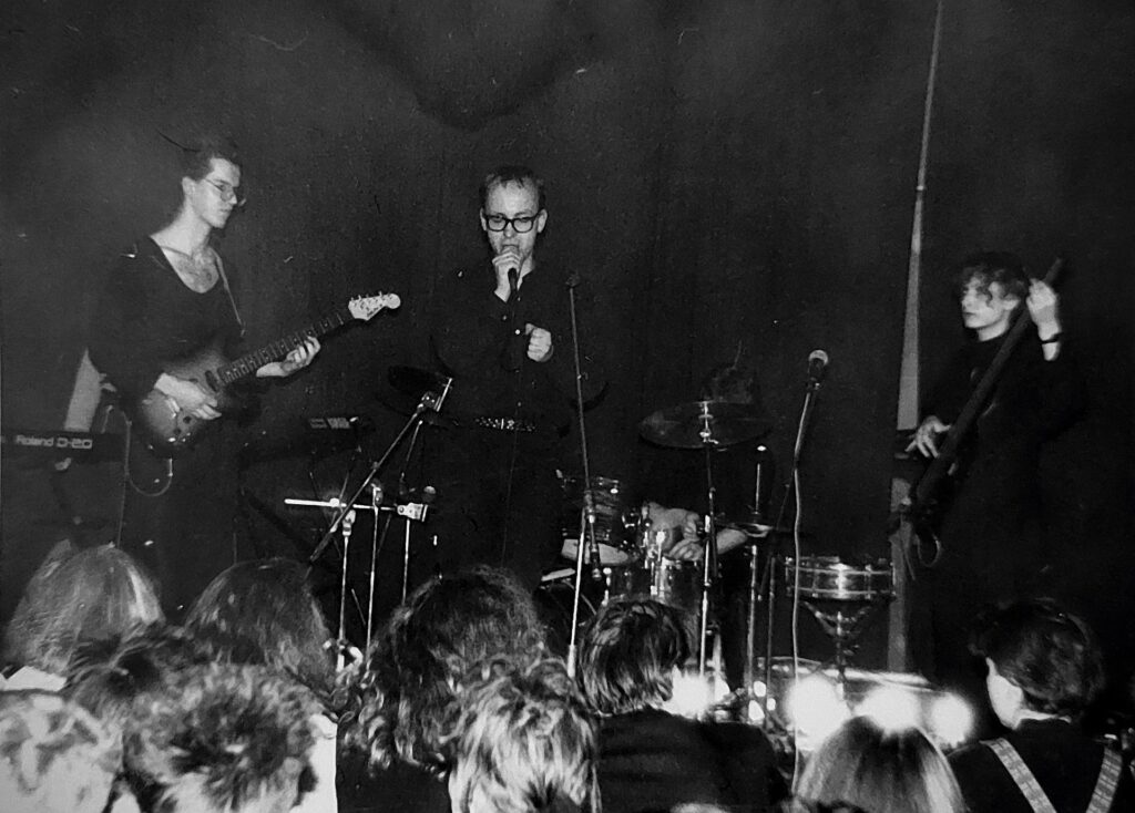 Rosengarten live auf einer dunklen Bühne, circa 1989 – Gitarrist, Sänger mit Brille und Bassist, Publikum von hinten sichtbar
